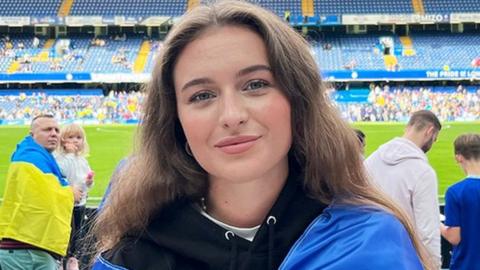 Marta, a Ukrainian woman with a blue and yellow Ukrainian flag around her looking at the camera smiling. She is wearing a black hoody. The background is of Stamford Bridge football stadium, with other people dressed in Ukrainian colours, a green grass pitch and blue seats.