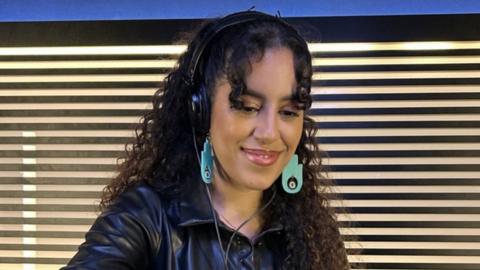 Nooriyah smiles as she mixes in a recording studio. She's got shoulder-length brown curly hair, with large, dangly cyan-coloured earrings in a Hand of Fatima design. That's a flat with an eye in the middle of the palm. Nooriyah's also wearing over-ear headphones and a black leather jacket.