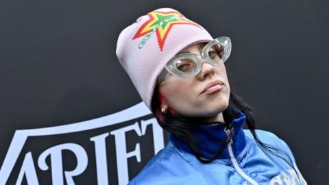 Billie Eilish attends the 2023 Variety Hitmakers Brunch at NYA WEST on December 02, 2023 in Hollywood. Billie, a 21-year-old woman, wears a pink beanie hat with a green, yellow and red star on the front. She wears thick rimmed clear glasses and a blue zip-front jacket. She's pictured in front of a black background with 'Variety' printed on it.