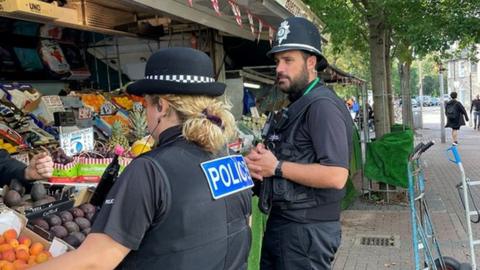 Two police officers, one male and the other female. They are standing next to a fruit stall. The officers are facing towards the stall.