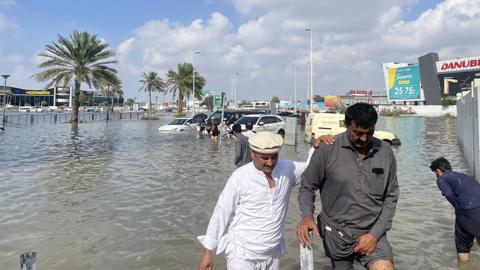 People wade through flood water with trees and clouds in the background, in Dubai on 17 April
