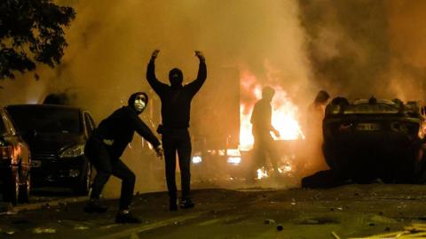 Hooded people standing with arms raised in front of burning cars