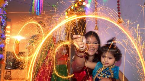 Family celebrating Diwali with firecrackers. A woman and child are the focus, holding sparklers, waving it in a circle creating a ring of orange sparks. The child is wearing a blue traditional dress. In the back is another woman and child also holding sparklers and waving in a circle creating orange sparks. There are also multi coloured fairy lights visible on the walls.