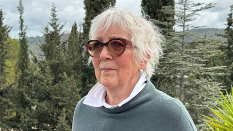 A white senior woman with white hair and coloured glasses looks into the distance in front of a backdrop of greenery