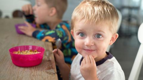 A young child sits at a table, on the table is a pink bowl with breakfast cereal in it. The child looks and smiles at the camera. Another child in the background is spooning food into their mouth.