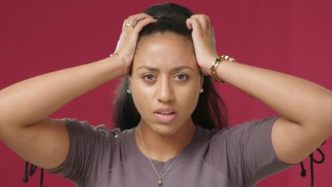 A teenage student holds her hands to her head in exasperation on a maroon background.