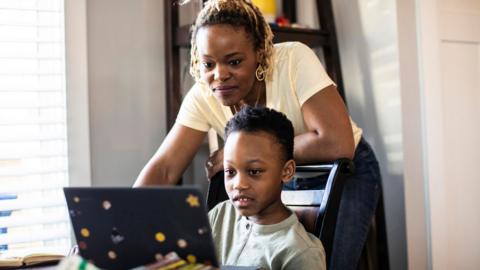 A young boy looks at a laptop while his mother smiles behind him. 