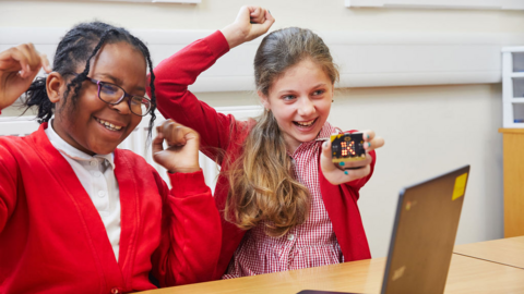 Two schoolchildren wearing uniform in a classroom hold up a micro:bit in celebration.