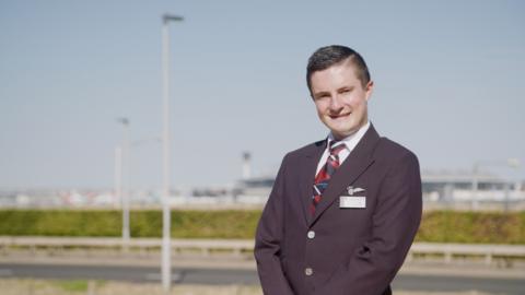 A member of air cabin crew stands in uniform outside an airfield.