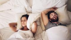 A woman covers her ears with a pillow while a man snores while sleeping