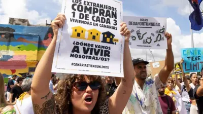 Manifestante con una pancarta que dice: Los extranjeros compran 1 de cada 3 viviendas en Canarias. ¿Dónde vamos a vivir nosotras/os?