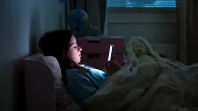 A young girl lies in bed in the dark, the light from a mobile phone in her hands lighting her face
