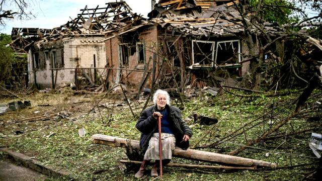 A woman sits on a fallen log in front of two houses that have been demolished by a missile strike, which killed another woman, in Druzhkivka, Donetsk