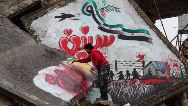 A man draws a slogan in Arabic reading "we still want freedom" as part of a graffiti mural commemorating the ninth anniversary of uprising that led to the Syrian conflict (March 15), showing a dove holding an olive branch in its beak flying over a Syrian opposition flag in the shape of the eastern Arabic numeral "9" while being targeted by the silhouette of a military aircraft with the Arabic word "years" below as silhouettes of children stand by a border fence and a tent with the letters "UN", drawn on the collapsed roof of a heavily damaged building in the town of Binnish in the northwestern Idlib province on March 13, 2020. (