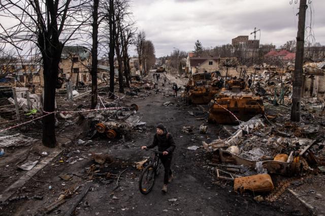 A man pushes his bike through debris and destroyed military vehicles on a street in Bucha, Ukraine - 6 April 2022.