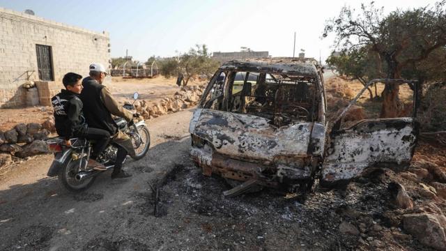 сирийцы на мотоцикле проезжают мимо обгоревшей машины