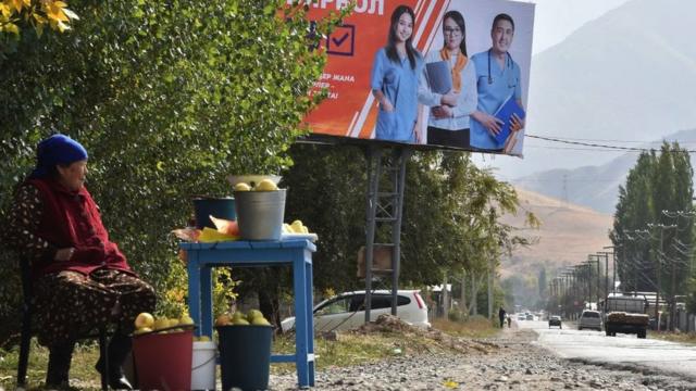Женщина продает яблоки у дороги на фоне предвыборного плаката