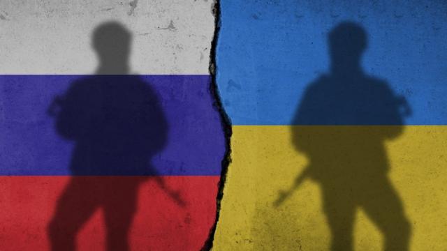 Two silhouettes of soldiers holding a gun stand in front of flags - to the left a Russian flag, to the right a Ukrainian flag
