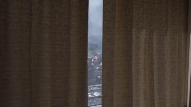 Через полузадернутые шторы виден дом, загоревшийся в результате обстрела, 12 марта 2022 года (фотография Мстислава Чернова)