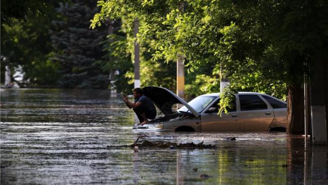 Человек сидит на открытом капоте автомашины посреди наводнения