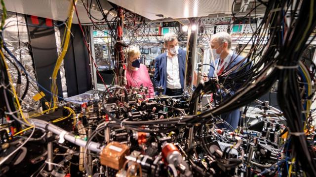 Angela Merkel stands next to two men in front of a complex, somewhat messy array of pipes and wires