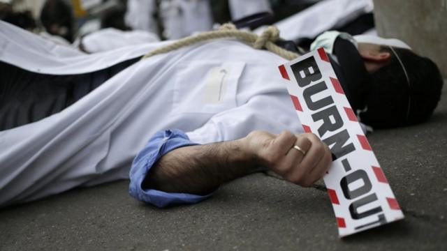 Врач в медицинском халате и с петлей на шее держит табличку с надписью "выгорание" во время акции возле здания Министерства здравоохранения Франции в Париже, февраль 2014 года.