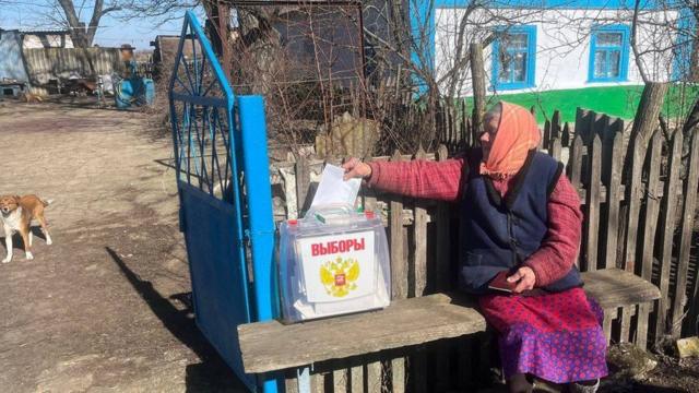 Бабушка опускает бюллетень в урну, сидя на скамейке рядом с домом