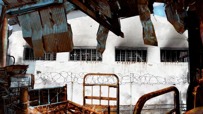 A white prison building from Olenivka framed by the destroyed interior of the warehouse hit by explosions.