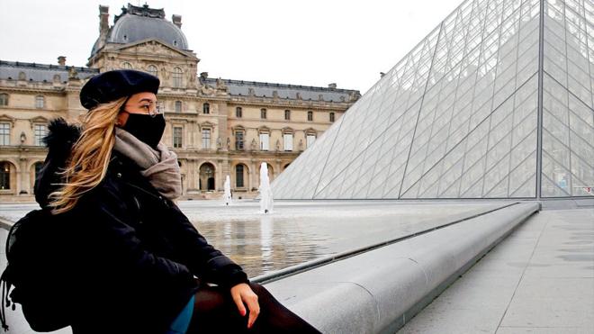 A women with a mask sitting in front of Louvre in Paris