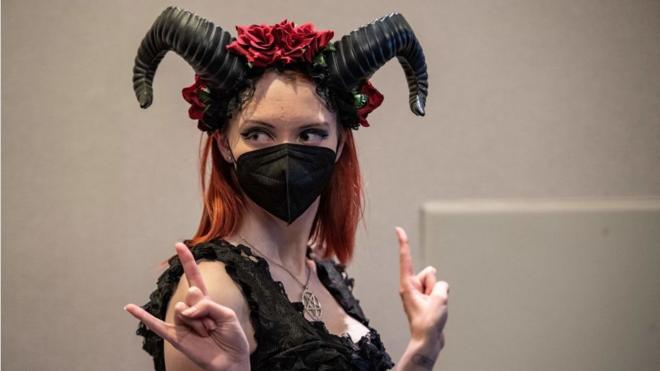 A woman wearing elaborate black horns and a Covid face mask poses for photos giving the sign of the horns at SatanCon in Boston, Massachusetts