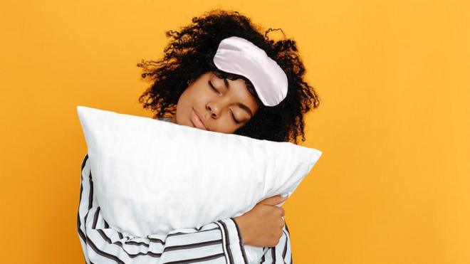 Woman hugging a white pillow, sleeping mask over her head, bright yellow background