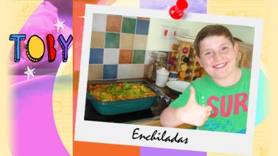 A little boy (Toby) grinning next to a stove top showing a dish of enchiladas.