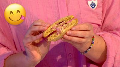 A womans hands are holding a cookie which has been broken in half. Inside the cookie is a strawberry cheesecake filling. 