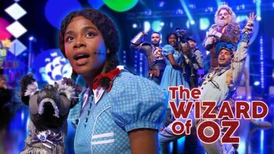 Dorothy from The Wizard of Oz theatre production, she is a black woman with hair tied into pig tails with a red ribbon, she is singing with her dog Toto who is a puppet, the rest of the cast are posing in the background.