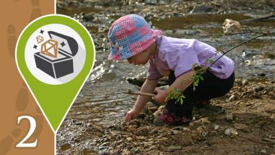 Little girl collecting objects from a muddy patch next to a stream with a treasure chest graphic over the image