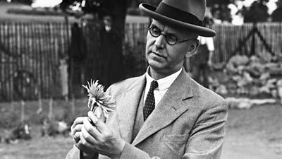 A man in a tweed suit and hat examines a flower