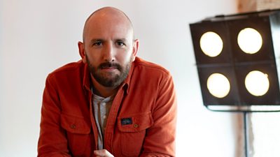 David Carlyle looking to camera slightly leaning forward. studio lighting in the background