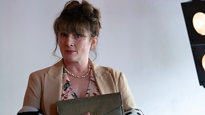 Sally Howitt looking to camera clutching a small handbag. Studio light in background