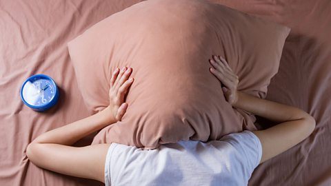 Person lying in bed with pillow over their head frustrated at their inability to sleep