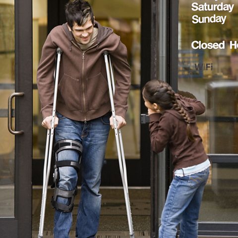 Young girl with jeans and a brown jumper and a poneytail is holding a glass door for a man on crutches with a knee brace also wearing jeans and a brown jumper.