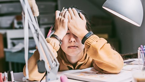 Teenage girl holds her head in her hands at a desk as she tries to concentrate on her revision or studies