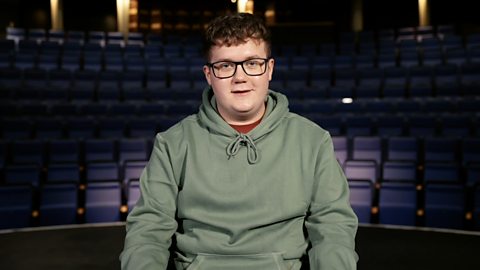 A student with glasses wearing a green hoodie looks at the camera, sitting in front of a lecture hall.