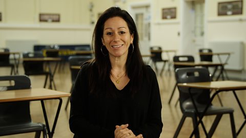 Dr Anna Colton smiling wearing a black shirt and sitting in front of an exam hall, with brown exam desks and black chairs behind her.