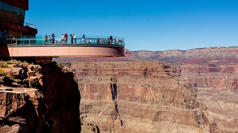 Photo of the Grand Canyon Skywalk extending out into the canyon. The bottom of the structure is made from orange-brown steel beams in a U shape, with a glass barrier above.