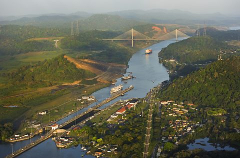 A photo of a lock on the Panama Canal. In the distance there is a white bridge across the river, and either side are forest-covered hills. On the river there are several ships.