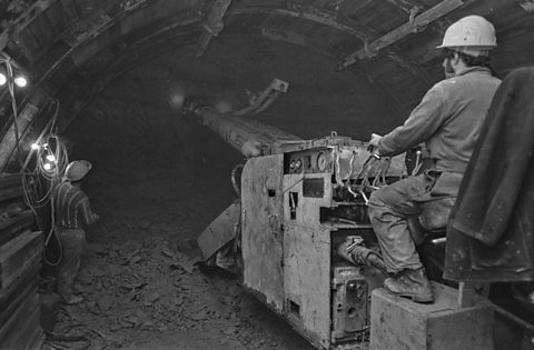 A black and white photograph of a male construction worker using machinery to bore a hole for the Channel Tunnel. The floor is covered with bits of rock.