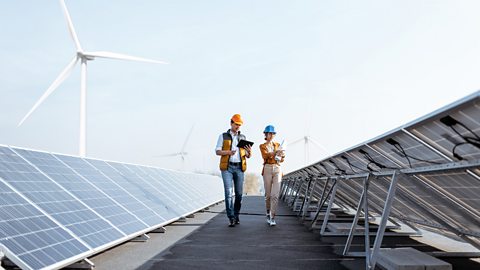 Two workers in hard hats inspect rows of solar panels, with wind turbines in the background