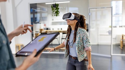 Woman wearing a VR headset in the office looks around in wonder, while another person operates the software from a tablet.