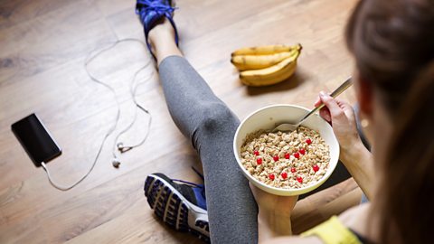 A person sits on the floor in sports wear eating porridge with bananas nearby