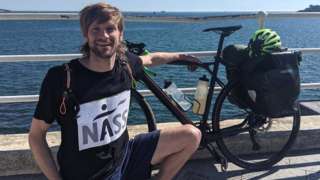 Man next to a body of water with a bike, wearing a shirt that reads NASS